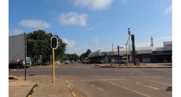 A quiet urban intersection with buildings, trees, and a clear blue sky. Traffic lights and a few vehicles are present, creating a calm, sunny morning scene.