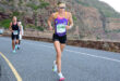 A runner in a purple tank top and black shorts races on a mountain road with determination. Other runners follow. The rocky backdrop suggests a challenging terrain.