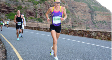 A runner in a purple tank top and black shorts races on a mountain road with determination. Other runners follow. The rocky backdrop suggests a challenging terrain.