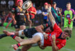 Two rugby players in action, one in a black uniform tackling another in a red uniform mid-air. The focus is intense, crowd and officials in the background.
