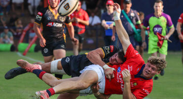 Two rugby players in action, one in a black uniform tackling another in a red uniform mid-air. The focus is intense, crowd and officials in the background.