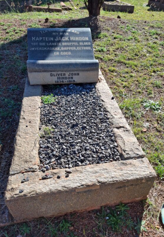 A stone grave with an engraved headstone sits in a grassy area. The text mentions "Kaptein Jack Hindon" and "Oliver John Hindon, 1874-1919." Stones fill the rectangular area, evoking a solemn mood.