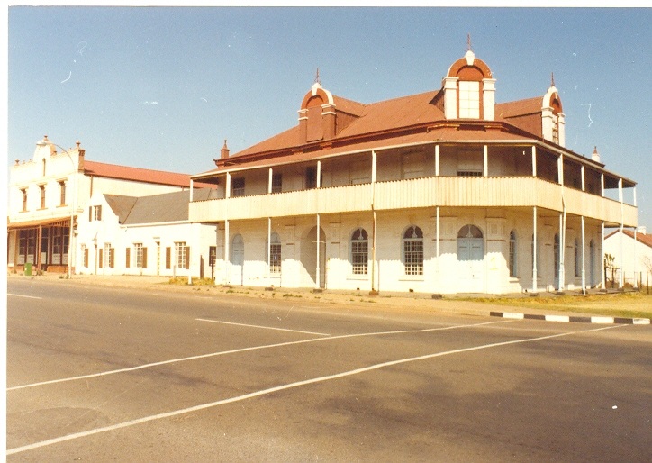 Historic building with Victorian architecture and red roofs in a sunny, deserted street. The structure features arched windows and a wraparound balcony.