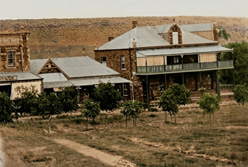 Historic stone buildings with metal roofs and a balcony stand amid sparse greenery on a rural landscape, conveying a sense of rustic serenity.