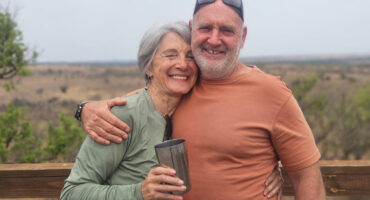 Smiling older couple embracing on a wooden deck overlooking a serene landscape; the woman holds a metal cup. Warm, joyful, and relaxed atmosphere.