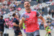 Rugby player in a pink jersey celebrates on a field, fist raised, expressing joy. Blurred spectators and players are visible in the background.