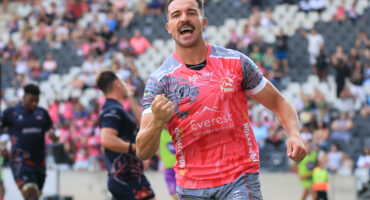 Rugby player in a pink jersey celebrates on a field, fist raised, expressing joy. Blurred spectators and players are visible in the background.