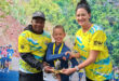 A young boy receives a trophy from a smiling man and woman in matching athletic shirts, set against a vibrant backdrop of foliage and runners.