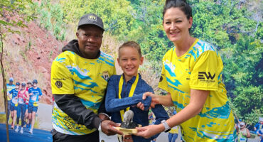 A young boy receives a trophy from a smiling man and woman in matching athletic shirts, set against a vibrant backdrop of foliage and runners.