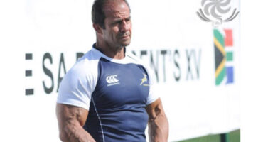A muscular man in a navy and white rugby shirt holds a Tbilisi Cup 2013 rugby ball, standing confidently. A "SARRACENS XV" sign and flag in the background.