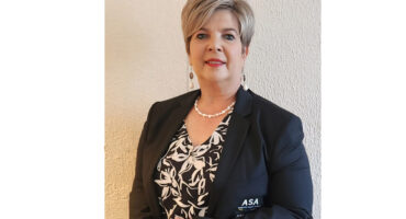 A confident woman stands against a cream wall, wearing a black blazer and floral blouse. She has short, styled hair, pearl necklace, and earrings.