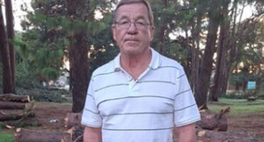 A man wearing a striped polo shirt stands in a wooded area with cut logs in the background. The setting is serene with tall trees and soft lighting.