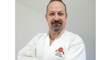 Man in a white martial arts uniform with folded arms smiles confidently. His attire has a logo and "South Africa" text, suggesting an official affiliation.