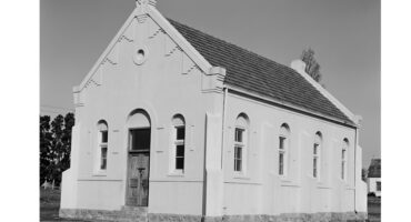 A small, white, historical church with arched windows and a wooden door, set against a clear sky. The stone foundation suggests an old, serene atmosphere.