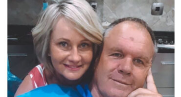 A smiling couple poses in a kitchen with tiled walls. The woman has short, blonde hair and wears a striped top. The man rests his head on his hand.