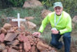 An elderly person in bright clothing kneels beside a pile of rocks with a white cross in a lush, green outdoor setting, conveying a somber yet reflective mood.