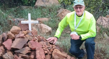 An elderly person in bright clothing kneels beside a pile of rocks with a white cross in a lush, green outdoor setting, conveying a somber yet reflective mood.