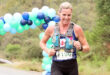 A woman running a marathon, wearing a hydration vest and bib number. She is near a colorful balloon arch, with a background of trees and a road.