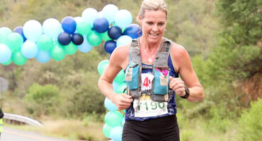 A woman running a marathon, wearing a hydration vest and bib number. She is near a colorful balloon arch, with a background of trees and a road.