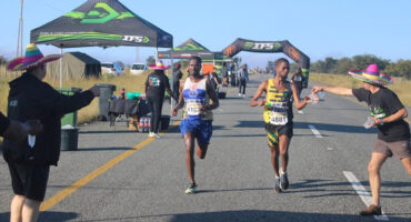 Two marathon runners in race bibs approach a hydration station on a sunny day. Volunteers in sombreros offer drinks, creating a festive atmosphere.