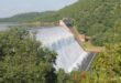 A large dam overflows with water cascading down its concrete slope. Surrounded by lush green hills and foliage under a clear blue sky.