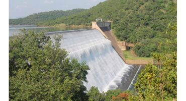 A large dam overflows with water cascading down its concrete slope. Surrounded by lush green hills and foliage under a clear blue sky.