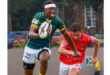 A focused rugby player in a green and yellow jersey runs with the ball, pursued by an opponent in a red jersey. The scene is dynamic and intense.