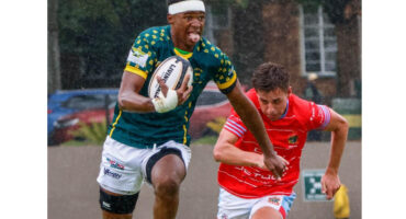 A focused rugby player in a green and yellow jersey runs with the ball, pursued by an opponent in a red jersey. The scene is dynamic and intense.