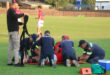A group of medics tends to an injured rugby player lying on a the field. The medics are focused and preparing to lift the player onto a stretcher.