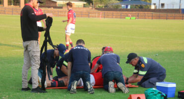 A group of medics tends to an injured rugby player lying on a the field. The medics are focused and preparing to lift the player onto a stretcher.