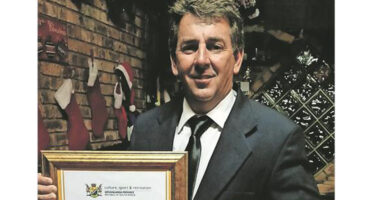 A smiling man in a suit holds a framed certificate in front of a brick wall decorated with Christmas stockings. The atmosphere is festive and celebratory.