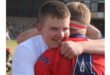 Two young men embrace on a sports field, one in a white shirt and the other in a red jersey. Their expressions convey joy and camaraderie.