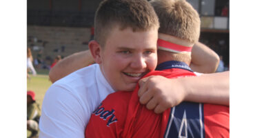 Two young men embrace on a sports field, one in a white shirt and the other in a red jersey. Their expressions convey joy and camaraderie.
