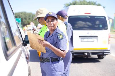 A police officer in uniform holds a clipboard, standing by a stopped vehicle on a sunny day. Two other officers are visible in the background.
