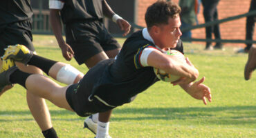 A rugby player in a black jersey dives across the grass, holding the ball tightly. Teammates surround him, capturing a moment of action and focus.