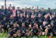 A jubilant rugby team poses for a group photo on a grassy field. Players in black jerseys cheer with raised fists, exuding joy and team spirit under a cloudy sky.