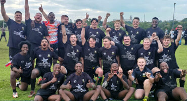 A jubilant rugby team poses for a group photo on a grassy field. Players in black jerseys cheer with raised fists, exuding joy and team spirit under a cloudy sky.