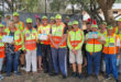 A diverse group of people in safety vests and caps pose outdoors under a tree. Some hold easter eggs, creating a sense of community and teamwork.