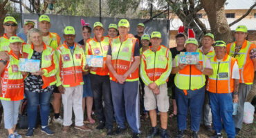 A diverse group of people in safety vests and caps pose outdoors under a tree. Some hold easter eggs, creating a sense of community and teamwork.