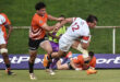 Rugby match action: A player in orange grasps an opponent in white, numbered 12, mid-air. Other players and green field visible, conveying intensity.