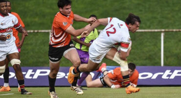 Rugby match action: A player in orange grasps an opponent in white, numbered 12, mid-air. Other players and green field visible, conveying intensity.