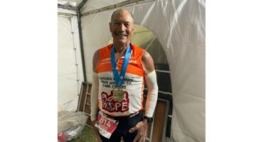 Elderly man smiling proudly with a medal around his neck, wearing a running outfit in front of a white tent. The atmosphere is joyful and triumphant.