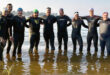Group of ten people wearing wetsuits and swim caps standing in knee-deep water, arms around each other, smiling at the camera. Background features a calm dam and trees.