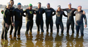 Group of ten people wearing wetsuits and swim caps standing in knee-deep water, arms around each other, smiling at the camera. Background features a calm dam and trees.