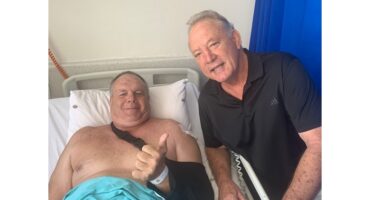 An older man with a bandaged arm gives a thumbs-up while lying in a hospital bed, smiling. Another man stands beside him, smiling supportively.