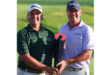 Two men wearing golf shirts and caps, smiling and holding a black and red trophy on a green golf course. The mood is celebratory and proud.