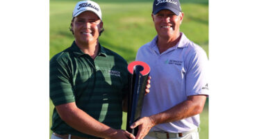 Two men wearing golf shirts and caps, smiling and holding a black and red trophy on a green golf course. The mood is celebratory and proud.