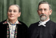 A man and woman wearing glasses pose against a green backdrop. The woman wears floral embroidery; the man has a bow tie. Both convey a calm, serious demeanor.