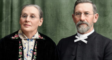 A man and woman wearing glasses pose against a green backdrop. The woman wears floral embroidery; the man has a bow tie. Both convey a calm, serious demeanor.
