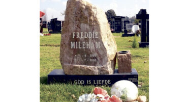 A tall, rustic gravestone with "Freddie Mileham" inscribed stands on a grassy cemetery. Flowers at the base. The tone is peaceful and solemn.